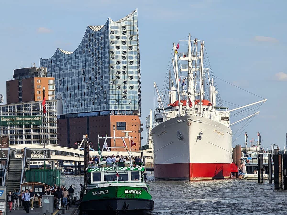 Blick auf die Elbphilharmonie mit ihrer modernen Glasfassade und das weiße, historische Segelschiff Rickmer Rickmers, das im Hamburger Hafen liegt. 
