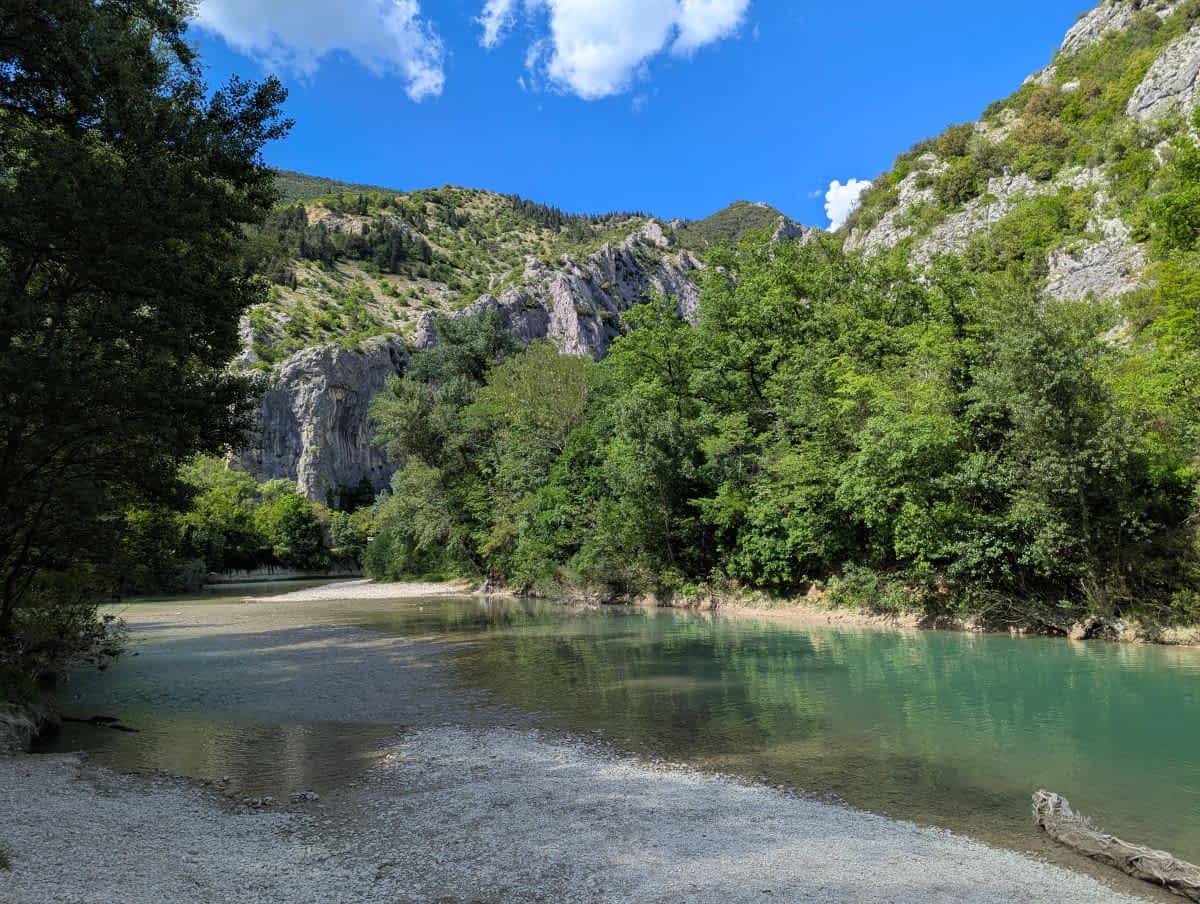 Türkisblaues Flusswasser und ein Kiesufer in der Furlo-Schlucht, umgeben von grünen Bäumen und felsigen Bergen