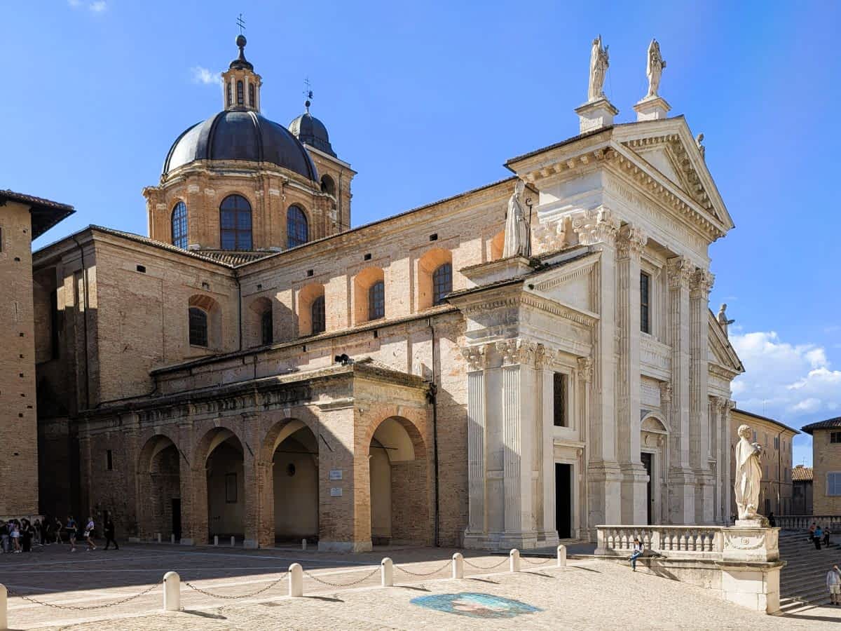 Klassizistische Fassade des Doms von Urbino mit Kuppel und Statuen unter blauem Himmel.