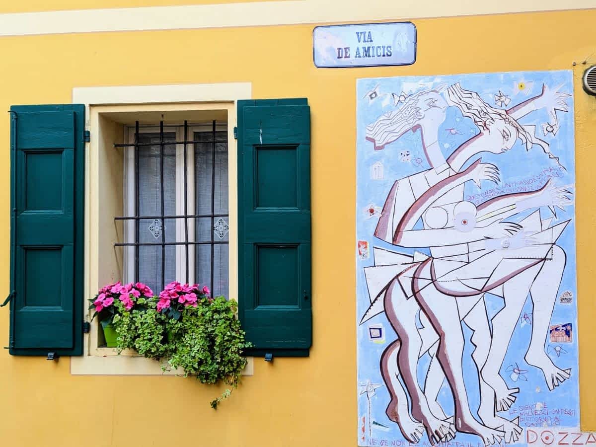 Gelbe Hauswand in Dozza mit einem abstrakten Wandgemälde in Blau und Weiß neben einem Fenster mit grünen Fensterläden.