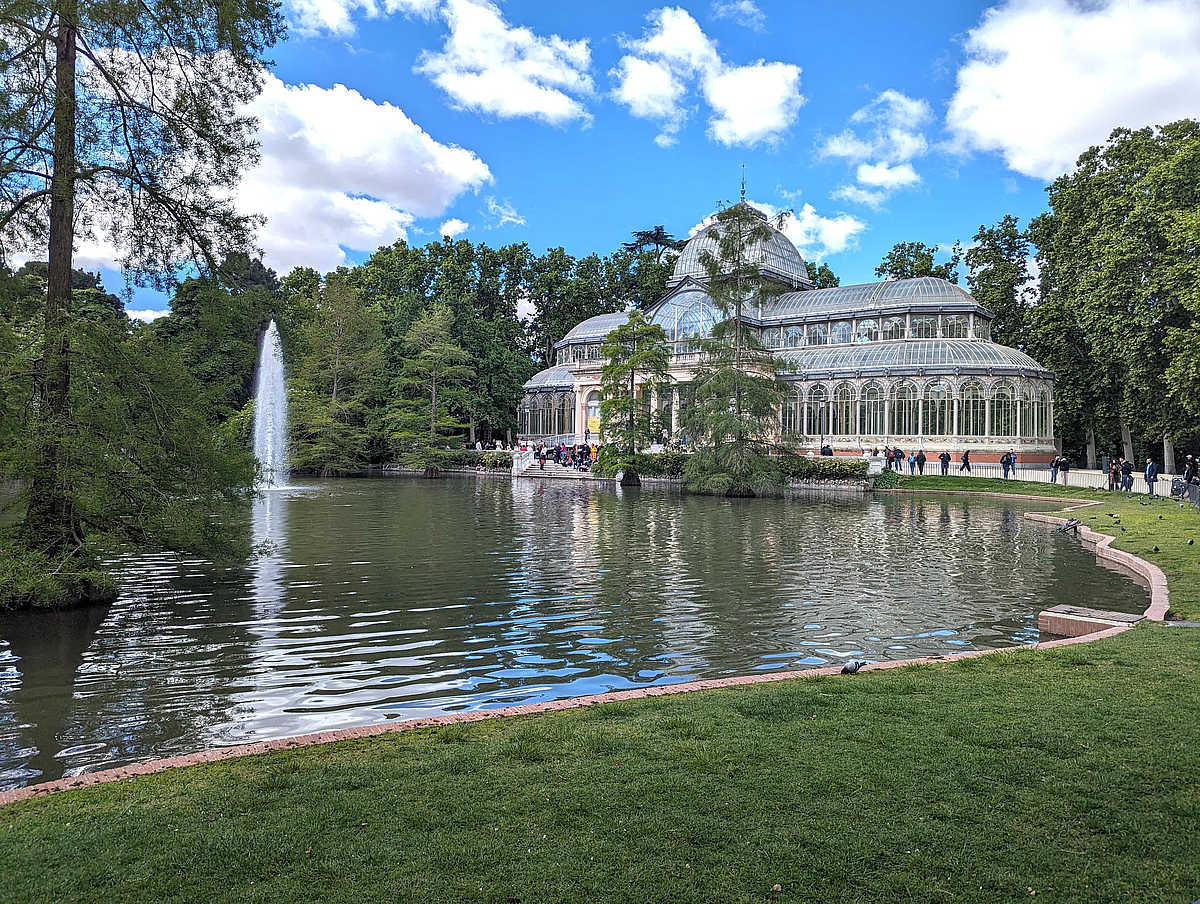 Glaspavillon mit filigraner Eisenkonstruktion im Grünen, reflektiert im Wasser.